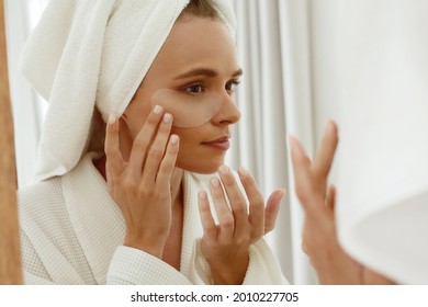 European girl applying under eye patch on face and watch in mirror. Young concentrated beautiful woman wear bathrobe and wrapped bath towel on head. Concept of face skin care - Powered by Shutterstock