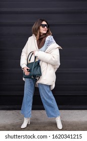European Girl In A Beige Oversized Down Jacket, Knitted Sweater, Jeans Flared With A Handbag And Glasses Is Standing On The Street. Life Style