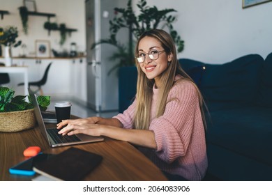 European Girl In Glasses Typing On Laptop Computer At Home. Concept Of Domestic Lifestyle. Idea Of Home Rest And Leisure. Young Beautiful Smiling Woman Sitting At Table. Interior Of Studio Apartment