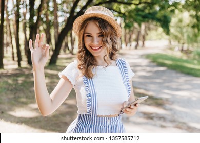 European girl in hat posing in park with happy face expression and waving hand. Joyful female model in vintage summer attire having fun.