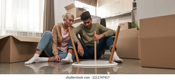 European Girl Looking At Her Middle Eastern Husband Or Boyfriend Assembling Table On Floor. Young Multiracial Couple Moving To New Apartment. Concept Of Home Relocation. Interior Of Spacious Flat