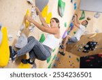 European girl is mastering climbing on training wall in gym, side view. Young woman holds on tightly to ledges and strives for top of bouldering route