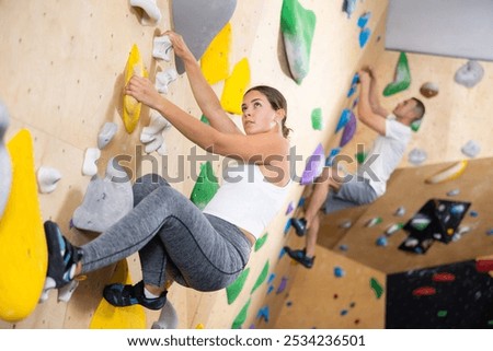 European girl is mastering climbing on training wall in gym, side view. Young woman holds on tightly to ledges and strives for top of bouldering route
