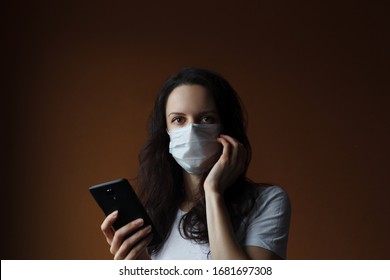 European Girl In Protective Medicine Mask With One Hand In Front Of Her Face And Another Hand Holding Cell Phone. News About Coronavirus Or Covid 19 Pandemic