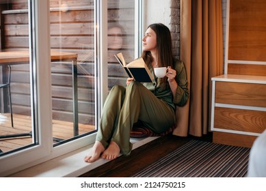 European Girl Reading Book And Drinking Tea Or Coffee At Home. Young Beautiful Woman With Brown Hair Wear Pajamas. Concept Of Rest. Modern Apartment. Daytime