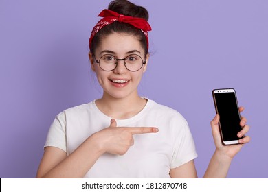 European girl showing blank cellphone screen with fore finger and looking directly at camera with charming smile, female with white casual t shirt and red hairband, lady with positive expression. - Powered by Shutterstock