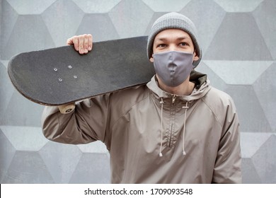 European guy in medical mask holds skateboard on his shoulder. Gray concrete wall background. Skateboarder in a gray jacket and hat. Coronavirus protection measures. - Powered by Shutterstock