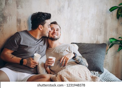 European happy gay couple drinking tea while having fun in bed , caressing and watching movies. Homosexual relationships and alternative love lifestyle concept