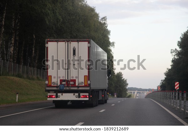 European heavy truck with white semi trailer\
van drive on left side of two lane suburban asphalted one way road,\
rear view at summer evening on forest and sky background,\
transportation\
logistics