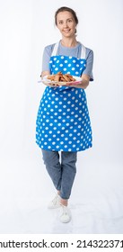 european housewife in a blue kitchen apron, jeans on a white background, full length portrait. a woman holds a white plate with cookies and points her finger or hand to the sides. place for writing