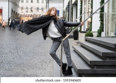 European lively young girl in gray textile pants, an oversized warm leather jacket, nubuck uggs and a large scarf actively posing on steps near store. Life style