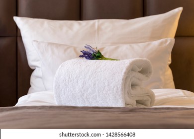 European luxury hotel bed detail with comfortable pillow in the back, and a decorative artificial flower on a rolled-up towel, prepared to greet and welcome guests.