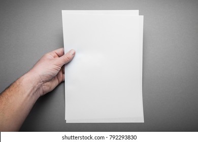 A European Male Hand Holds A Blank Sheet Of A4 Size Paper, A Newspaper On A Gray Background. Mockup.