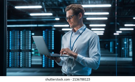 European Male IT Specialist Using Laptop Computer, Standing In Big Warehouse Data Center. System Administrator Working With SAAS. E-Business Digital Entrepreneur Examining Cloud Web Services