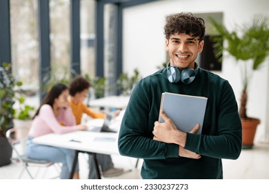 European man college student holding books and smiling at camera while female classmates studying with laptop on the background, coworking space - Powered by Shutterstock