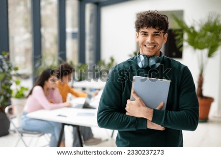 European man college student holding books and smiling at camera while female classmates studying with laptop on the background, coworking space