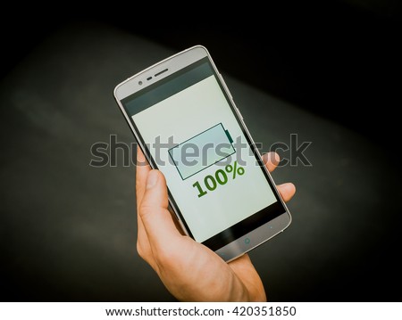European mans hand holding new silver smartphone on the black background. Full charge is on the screen,
