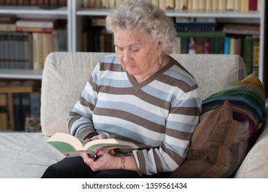 European Old Lady Reads Without Glasses At Home Against Bookshelves.