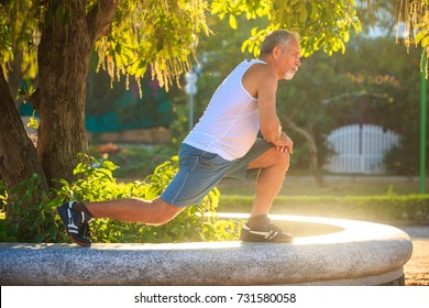 European Old Man In White Vest Blue Shorts Does Morning Exercises Squats On Left Knee On Stone Barrier By Tree