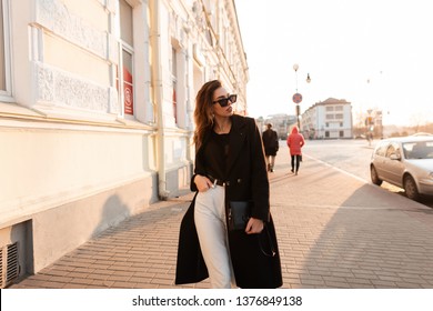 European pretty young hipster woman in a trendy black long coat in white jeans in a t-shirt posing standing in the city near the vintage white buildings. Stylish girl model enjoys the sunshine. 