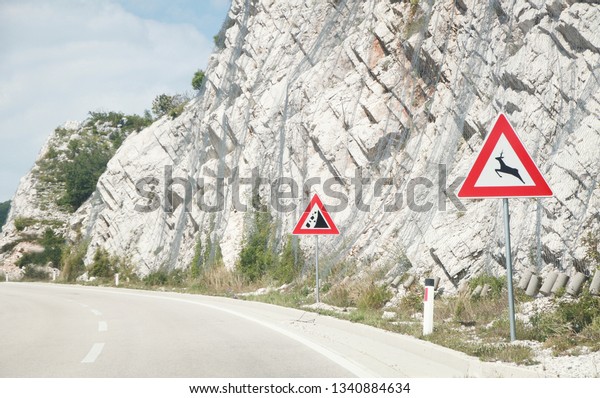 European road caution sign deer crossing\
by the roadway through plain countryside .Road sign. Stone-fall\
warning sign. Mountain road. Space for\
text.