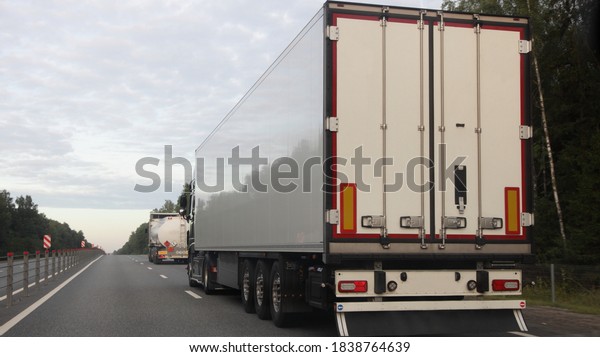 European road semi trucks with white trailer\
van and barrel move on two lane suburban asphalted highway\
motorway, rear view at summer evening on forest and sky background,\
delivery cargo\
logistics