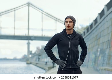 European runner standing at city quay. Young sportsman wearing hat, hooded zipper sweatshirt and mittens. Man prepare to sport run competition. Person listening music in earphones. Daytime - Powered by Shutterstock