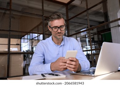 European smart businessman wearing glasses holding using cell mobile smartphone with laptop sitting at desk in office. Technology applications for business development growth and solutions concept.