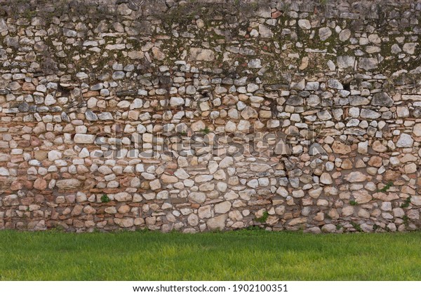 European stone wall constructed\
in warm tones, against edge of neatly trimmed green garden\
lawn