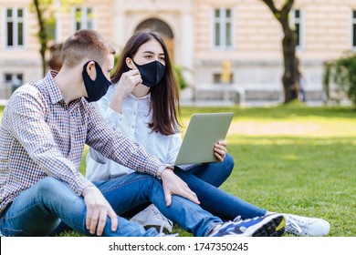 European Student Couple In Facial Masks Studying Outside On The Grass, Sitting Down On The School Campus Using Laptop Computer, Studying Hard Under The Trees Nature In Summer Sunlight.