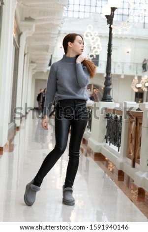 European tall girl with long red hair in black leather pants, gray winter boots and a fluffy sweater posing in a shopping gallery near boutiques