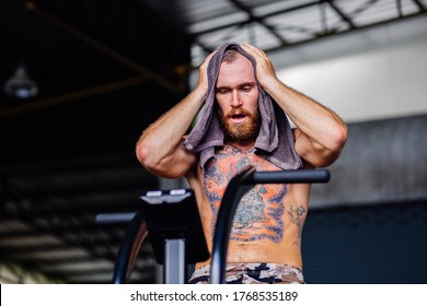 European Tattooed Bearded Exhausted Strong Tired Man Looks Sweaty After Doing Workout With Exercise Bike In The Gym Center, Clean Face With Towel, Sweating    