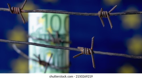 European Union Currency Wrapped In Barbed Wire Against Flag Of EU As Symbol Of Economic Warfare, Sanctions And Embargo Busting. Selective Focus On Barbed Wire