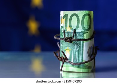 European Union Currency Wrapped In Barbed Wire Against Flag Of EU As Symbol Of Economic Warfare, Sanctions And Embargo Busting. Selective Focus On Barbed Wire. Copy Space.