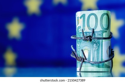 European Union Currency Wrapped In Barbed Wire Against Flag Of EU As Symbol Of Economic Warfare, Sanctions And Embargo Busting. Horisontal Image. Copy Space.