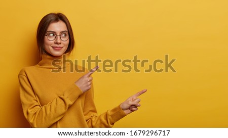 European woman with appealing appearance points fingers right, shows discount offer or banner, suggests visit link or webpage, wears turtleneck, isolated on yellow wall, gives recommendation.