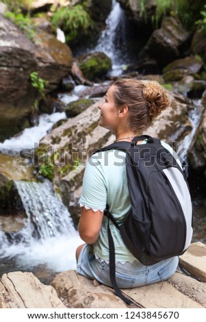European woman with backpack sits on stone and admires the beautiful views of mountain waterfall