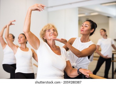 European woman doing ballet at a group training session in a dance studio performs an exercise near a ballet barre, where ..the choreographer helps to do it correctly