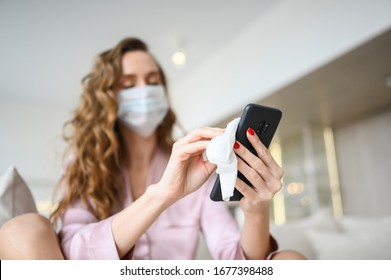 European woman in face mask cleaning the phone by hand sanitizer, using cotton wool with alcohol to wipe to avoid contaminating with Corona virus. Cleaning mobile phone to eliminate germs, Covid-19.  - Powered by Shutterstock