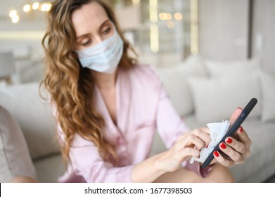 European Woman In Face Mask Cleaning The Phone By Hand Sanitizer, Using Cotton Wool With Alcohol To Wipe To Avoid Contaminating With Corona Virus. Cleaning Mobile Phone To Eliminate Germs, Covid-19. 