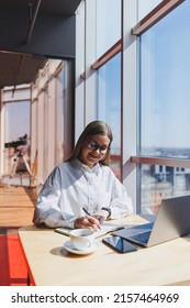 European Woman Manager Sits At A Table While Working On A Laptop And Drinks Coffee In A Cafe. A Young Smiling Woman Wears Glasses And A White Shirt. Freelance And Remote Work. Modern Female Lifestyle