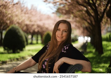 European woman sits on a park bench against a backdrop of an alley of ornamental cherry trees in full bloom with delicate pink blossoms, lush green grass - Powered by Shutterstock