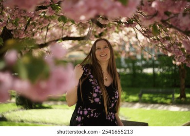 European woman smilingly admires the beauty of fully bloomed branches of delicate pink cherry blossoms overhead. Lush green foliage interspersed between.  - Powered by Shutterstock