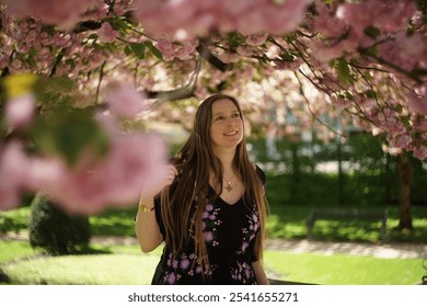 European woman smilingly admires the beauty of fully bloomed branches of delicate pink cherry blossoms overhead. Lush green foliage interspersed between.  - Powered by Shutterstock