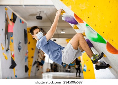 European young man in face mask grabbing ledges of artificial climbing wall in bouldering centre. - Powered by Shutterstock