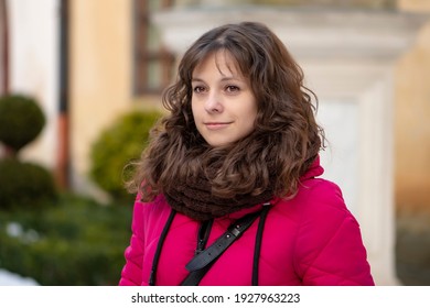 European Young Woman 30-35 Years Old With Dark Curly Hair Against The Backdrop Of A City Landscape With A Serious Look, Open Portrait.