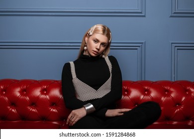 European Young Woman Fashion Model In Black Stylish Clothes With Trendy Pearl Hair Clips In A Fashionable Pearl Top Sits On A Leather Vintage Sofa In The Room. Glamorous Trendy Blonde Girl.Youth Style