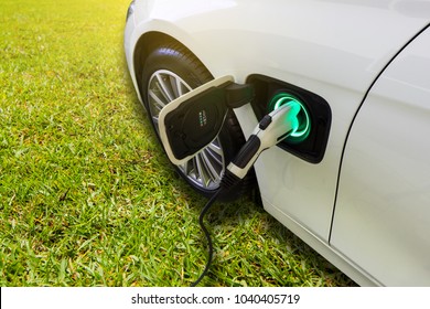 EV Car Or Electric Car At Charging Station With The Power Cable Supply Plugged In On Green Grass Nature With Sun Light Background. Eco-friendly Alternative Energy Concept