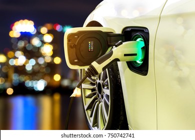 EV Car Or Electric Car At Charging Station With The Power Cable Supply Plugged In On Blurred Night Cityscape Background. 
