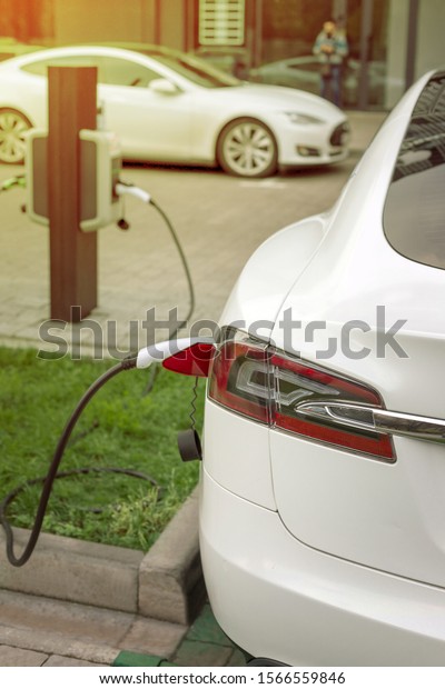 EV Car or Electric\
car at charging station with the power cable supply plugged in on\
blurred city background
